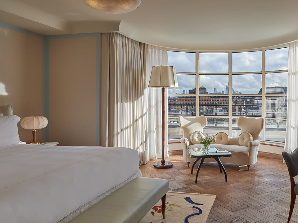 Elegant hotel suite with parquet flooring, a large bed in the foreground, and a curved wall of floor-to-ceiling windows revealing a city skyline. The sitting area features two sculptural white armchairs, a glass coffee table with fresh flowers, and a tall floor lamp beside sheer curtains.