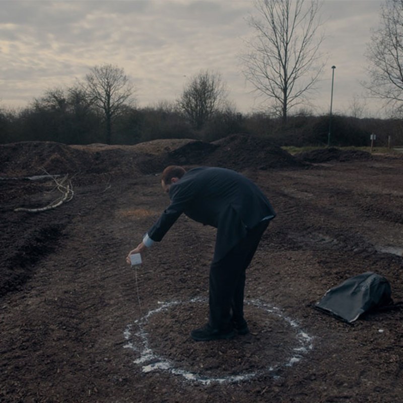 Man in a suit pouring white powder in a circle on the ground at an outdoor site.