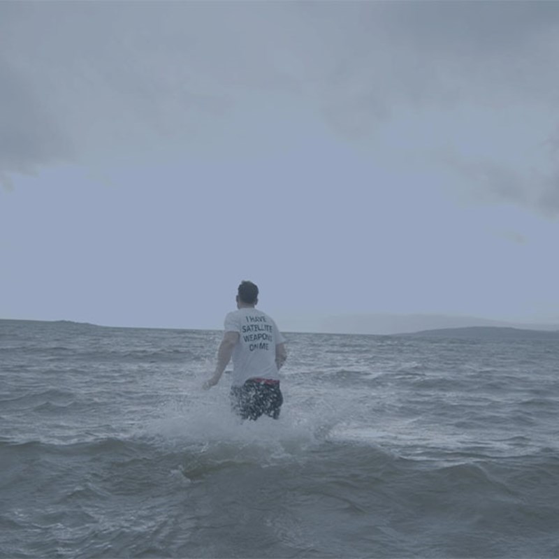 Person wearing a white T-shirt wading into the sea, with waves splashing around them under a cloudy sky.
