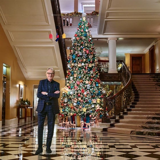 A man standing in front of a grand, ornately decorated Christmas tree in a luxurious, marble-floored staircase hall.