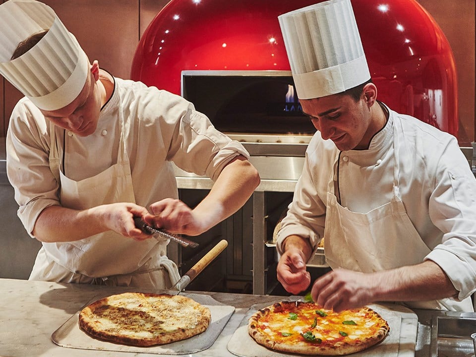 Photo of smiling and focused chefs preparing pizza and topping with parmesan cheese and garnish.