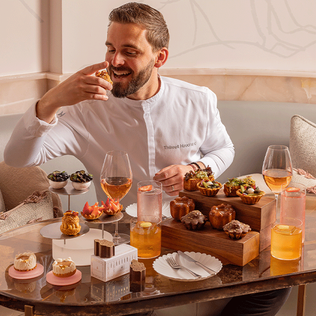 A smiling chef in a white uniform, seated at a marble table in a softly lit lounge, enjoys an elaborate afternoon tea spread with pastries, tarts, and drinks including rosé wine and cocktails.