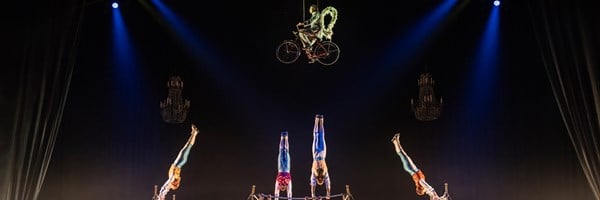 A panoramic view of an elaborate circus act showcasing synchronized acrobatics on bars and a performer gracefully cycling high above the stage under dramatic lighting.