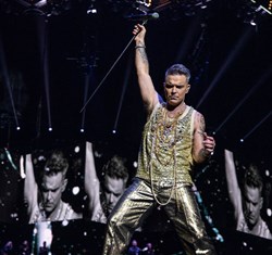 Robbie Williams performing, wearing gold and holding microphone and stand in the air, with screens around him projecting this