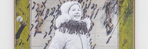 A mixed-media artwork featuring a black-and-white photograph of a smiling child in winter clothing, superimposed on a colorful aerial view of an urban plaza with people walking in various directions, casting long shadows.