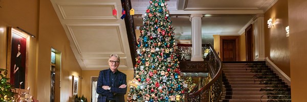 A man standing in front of a grand, ornately decorated Christmas tree in a luxurious, marble-floored staircase hall.