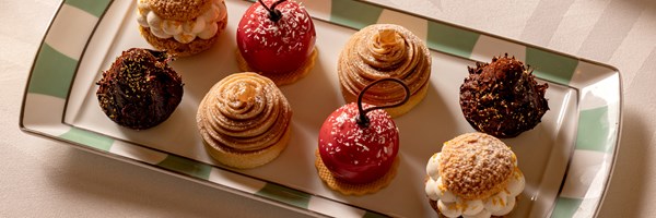 A close-up of an array of artfully crafted pastries, including glossy red confections, creamy swirls, and rich chocolate treats, elegantly presented on a rectangular platter with green and white accents.