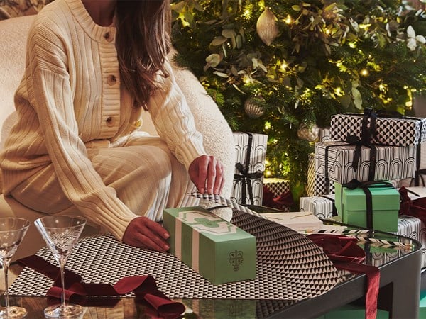 A cozy holiday scene with a woman in cream knit loungewear wrapping presents in black and white geometric paper, set against a beautifully decorated Christmas tree with twinkling lights and festive greenery.