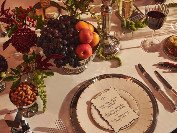 An opulent holiday dinner table arrangement, featuring a menu card atop fine china, a silver fruit bowl with grapes and peaches, fresh flowers, and delicate crystal glasses set for an indulgent meal.