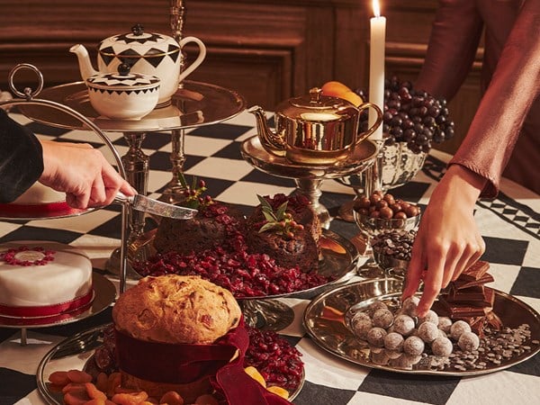 A lavish holiday dessert table adorned with rich Christmas pudding, a golden teapot, assorted chocolates, and dried fruits, set on a checkered tablecloth with vintage elegance.