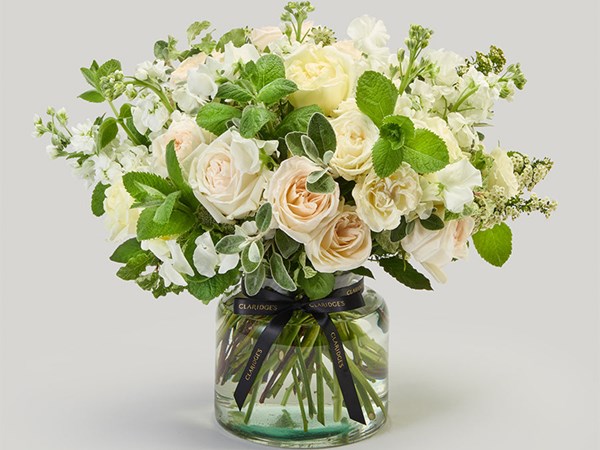 A sophisticated bouquet of white and ivory roses, accented with delicate green foliage and arranged in a glass vase tied with a black ribbon inscribed with 