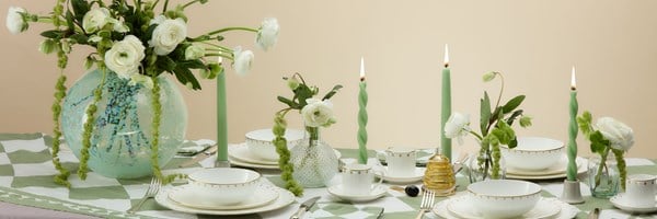 A panoramic view of an exquisitely styled dining table, showcasing white and gold china, green candles, and lush floral arrangements, with an artistic turquoise vase as the centerpiece.