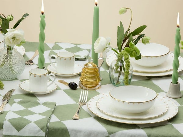 A sophisticated dining table set with fine white and gold-edged crockery, green twist candles, honey pot, and delicate floral arrangements atop a green and white geometric tablecloth.