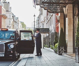 A smiling doorman opening the door of a taxi to welcome guests, outside the Claridge's Hotel.