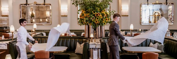 Inside a beautifully designed dining room, two staff members—one in a chef’s jacket and the other in a tailored suit—gracefully prepare the tables with a theatrical flourish of crisp white tablecloths.