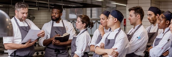 A head chef, dressed in his signature whites, leads a focused briefing in a professional kitchen, as an attentive team of chefs listens intently, taking notes.