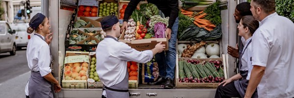 A group of chefs in crisp white uniforms and navy aprons gather around a delivery truck, receiving fresh, vibrant produce from a supplier on a bustling city street.