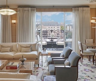 Elegant living area in Claridge’s Mews Pavilion Suite, featuring a neutral-toned interior with cream sofas, pale blue armchairs, and a patterned rug. A glass coffee table holds books and a decorative object. Floor-to-ceiling curtains frame large windows that open onto a balcony with a small black table and chairs, offering a view of classic Mayfair rooftops under a bright blue sky. A dining area with upholstered chairs and a modern chandelier is visible to the right.