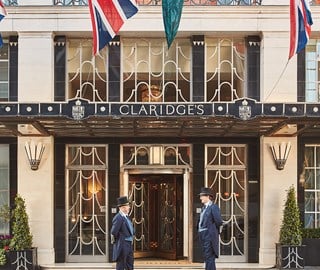 Two doormen in traditional uniforms and top hats stand outside the entrance of Claridge’s hotel in London, beneath the canopy with Union Jack flags above.