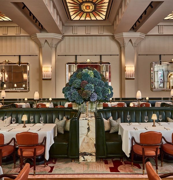 A grand dining room with opulent dark green leather booths, crisp white table settings, and a striking floral centerpiece, framed by ornate mirrors and stained-glass ceiling details.