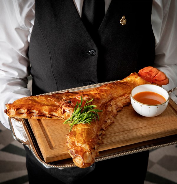 A waiter in an elegant black and white uniform presents a golden-brown lobster pie encased in delicate pastry, served on a silver tray with a side of rich bisque.