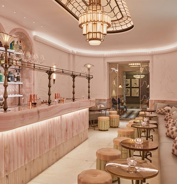 A softly lit, pastel-hued cocktail bar featuring a sculptural pink onyx counter, plush stools, and art deco-inspired chandeliers exuding an intimate, sophisticated ambiance.