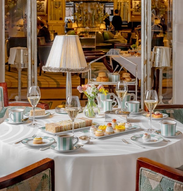 A refined afternoon tea setting with an elegantly dressed table featuring Claridge’s signature striped china, delicate finger sandwiches, exquisite pastries, and flutes of champagne, illuminated by a vintage table lamp in a grand, art deco-inspired dining space.