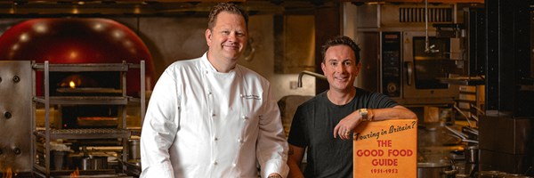 Two men are standing in a professional kitchen under a sign that reads 