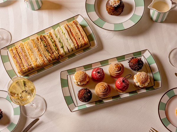 A refined afternoon tea display features an assortment of delicate finger sandwiches and intricately crafted pastries, including choux puffs, red-glazed confections, and Mont Blanc swirls, elegantly arranged on Claridge's signature green and white striped plates, accompanied by a glass of sparkling Champagne.