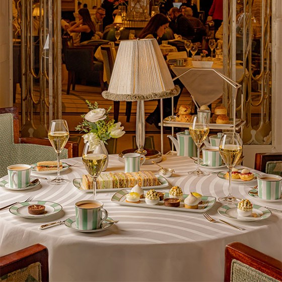 A beautifully set table at Claridge’s, showcasing an opulent afternoon tea spread with fine china, intricate pastries, finger sandwiches, and glasses of champagne, all under the warm glow of a fringed table lamp.