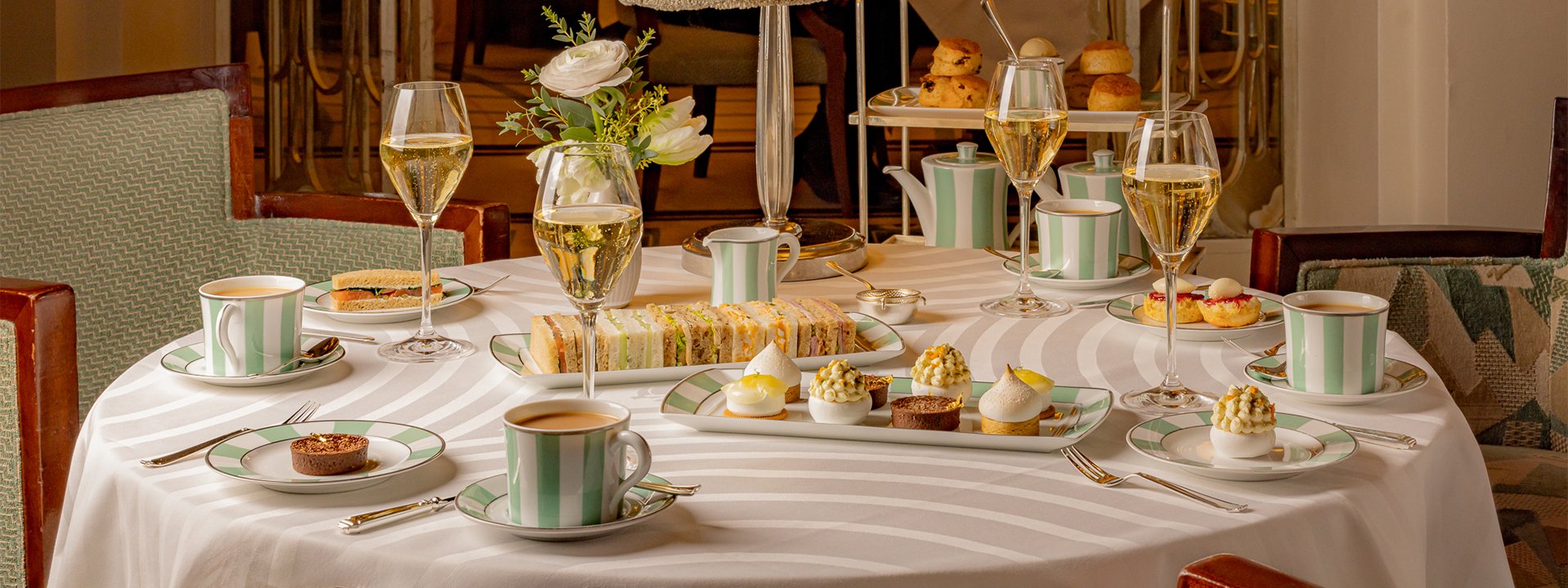 A grand afternoon tea setting at Claridge’s, elegantly arranged with delicate pastries, fine finger sandwiches, and signature green-and-white china, accompanied by sparkling champagne and freshly brewed tea, evoking a timeless sense of indulgence and refinement.