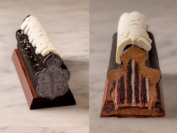 A luxurious chocolate yule log with intricate exterior detailing, topped with a white, textured garnish and sliced to reveal layers of chocolate sponge, cream, and berry filling.