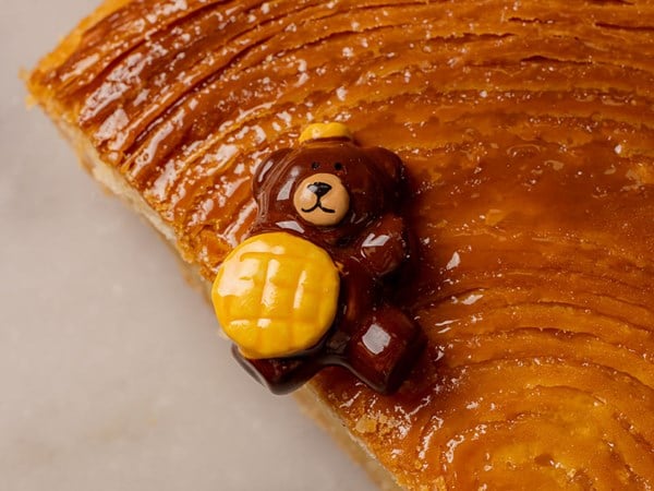 A golden-brown galette des rois with a glossy surface, adorned with a small chocolate bear holding a yellow honey pot.