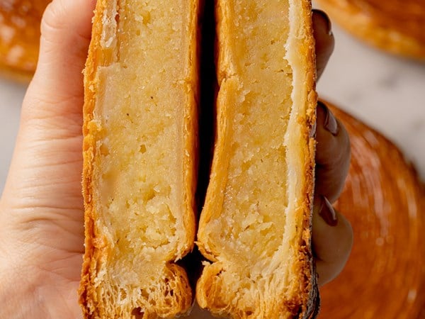 A close-up of a hand holding a sliced galette des rois, revealing flaky pastry layers and a creamy almond frangipane filling.