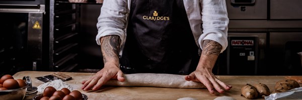 A baker with tattooed arms rolls dough on a floured work surface, surrounded by eggs, flour, and chocolate chip buns.