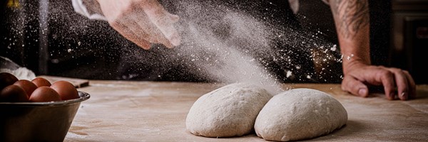 Flour is dramatically sprinkled over two proofing dough loaves, with eggs and baking tools nearby.