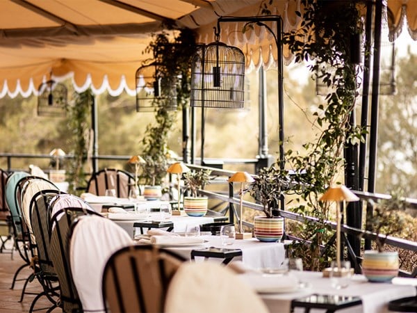 An inviting outdoor dining area under a striped canopy, adorned with potted plants, delicate hanging birdcage-style lamps, and colorful table accents, offering a relaxed yet sophisticated atmosphere.