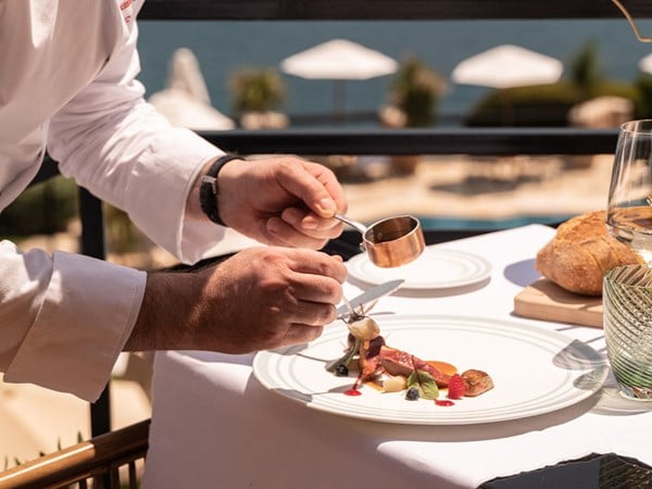 A chef artistically plating a gourmet dish on a terrace overlooking a serene seaside view, with bread, glassware, and pristine table settings completing the elegant dining experience.