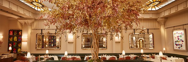 A festive dining room adorned with a dazzling golden tree centerpiece draped in metallic leaves and hanging stars, surrounded by elegant table settings and illuminated by stained-glass skylights.