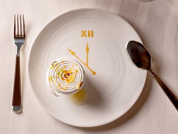 A delicate dessert adorned with gold leaf and citrus zest is elegantly presented on a clock-themed plate, accompanied by a glass of white wine and a vibrant red rose centerpiece on a pristine white tablecloth.