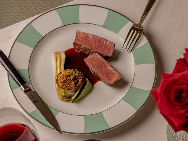 A refined plate featuring two slices of perfectly seared medium-rare beef, elegantly paired with a charred leek and a round of caramelized, herb-topped vegetable, all nestled in a rich, glossy sauce on a white plate with mint green and white accents.