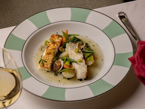 A sophisticated dish showcasing tender, pan-seared white fish accompanied by vibrant seasonal vegetables, delicate herbs, and a light broth, artfully presented on a white plate with mint green and white patterned borders.