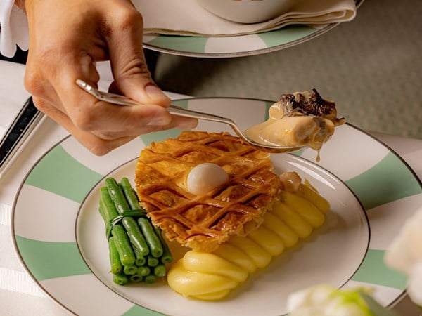 A delicate golden lattice-topped pastry filled with creamy, truffle-infused chicken, elegantly plated with piped mashed potatoes, a bundle of green beans, and a quail egg at its center, served on a green and white-rimmed plate.