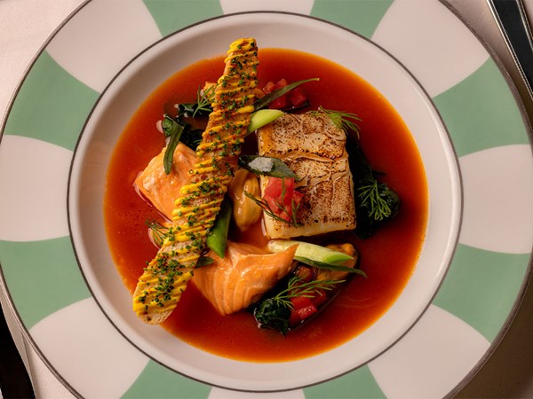 A refined seafood dish featuring seared cod and salmon in a rich, deep-red broth, garnished with fresh herbs, vegetables, and a crisp, herb-topped pastry twist, artfully presented on a signature green and white plate.