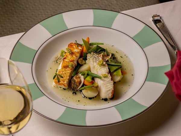 A beautifully plated fish dish with pan-seared fillet and shellfish, accompanied by vibrant spring vegetables in a light, aromatic broth, served on fine china with a glass of white wine nearby.