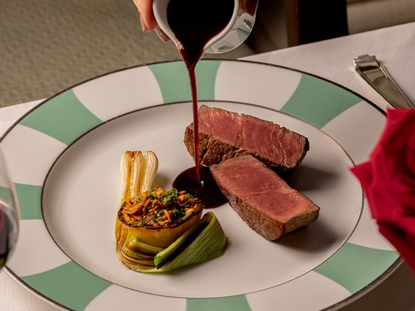 A luxurious cut of medium-rare beef, served with a roasted onion and charred leek, as a velvety red wine sauce is gracefully poured over, enhancing the dish’s rich flavors on an elegant, green-bordered plate.