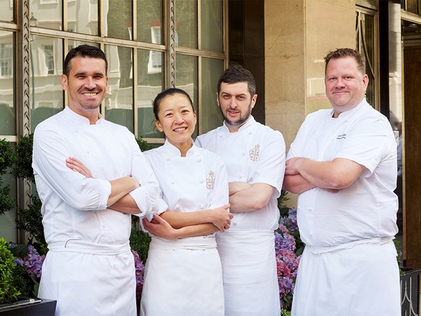 A group of four chefs, dressed in crisp white uniforms with embroidered logos, stand confidently outside a luxurious building with large windows and elegant stonework. They pose with arms crossed, smiling warmly, in front of a lush floral display, exuding a sense of teamwork and culinary excellence.