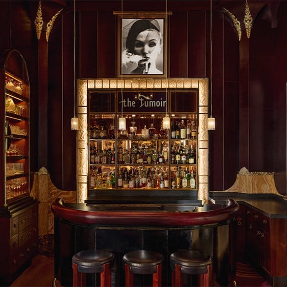 Interior view of The Fumoir bar at Claridge’s, showcasing a backlit Art Deco-style liquor display framed by carved golden panels, with pendant lights hanging over a glossy curved bar counter and richly textured dark walls