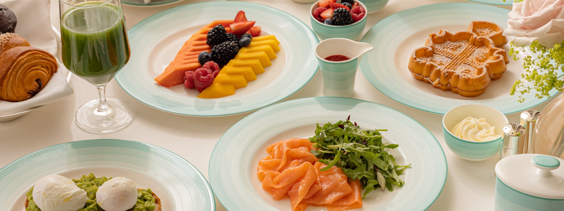 A close-up view of an elegant breakfast spread, featuring smoked salmon with arugula, fresh fruit, avocado toast with poached eggs, croissants, green juice, and a waffle, all served on pastel green and white dishware.