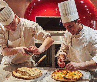 Photo of smiling and focused chefs preparing pizza and topping with parmesan cheese and garnish.